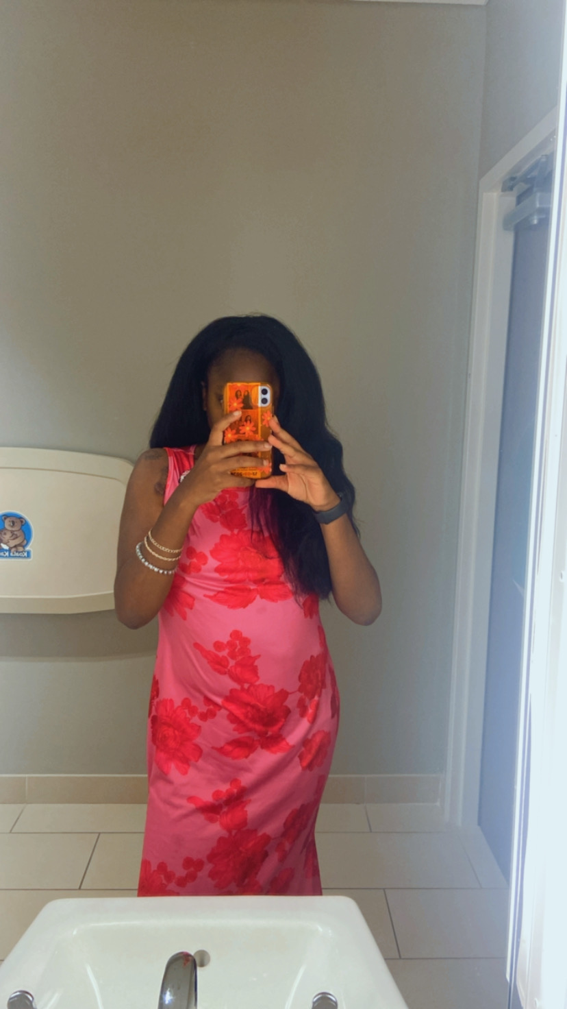 Person taking a mirror selfie in a pink floral dress, holding a phone with an orange case, standing in a room with beige walls and white furniture.