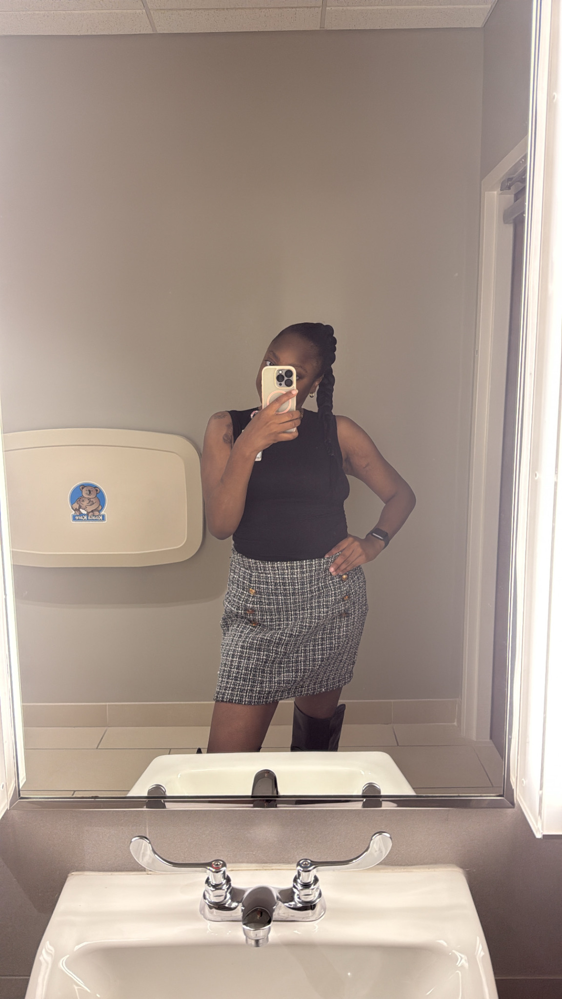 Person taking a mirror selfie in a bathroom, wearing a black top and patterned skirt, with a hand on hip.
