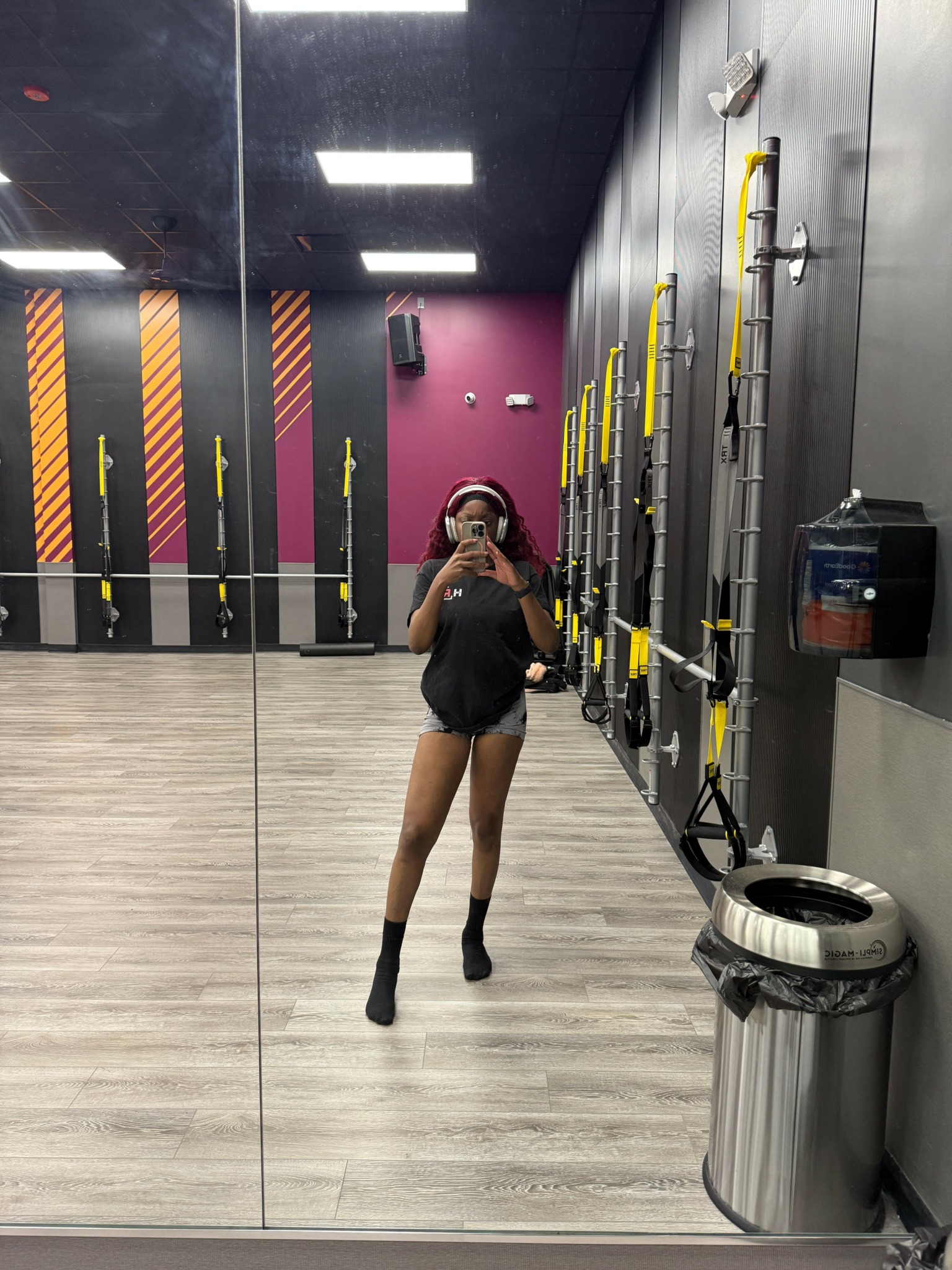 Person taking a mirror selfie in a gym, with fitness equipment and a pink wall in the background.