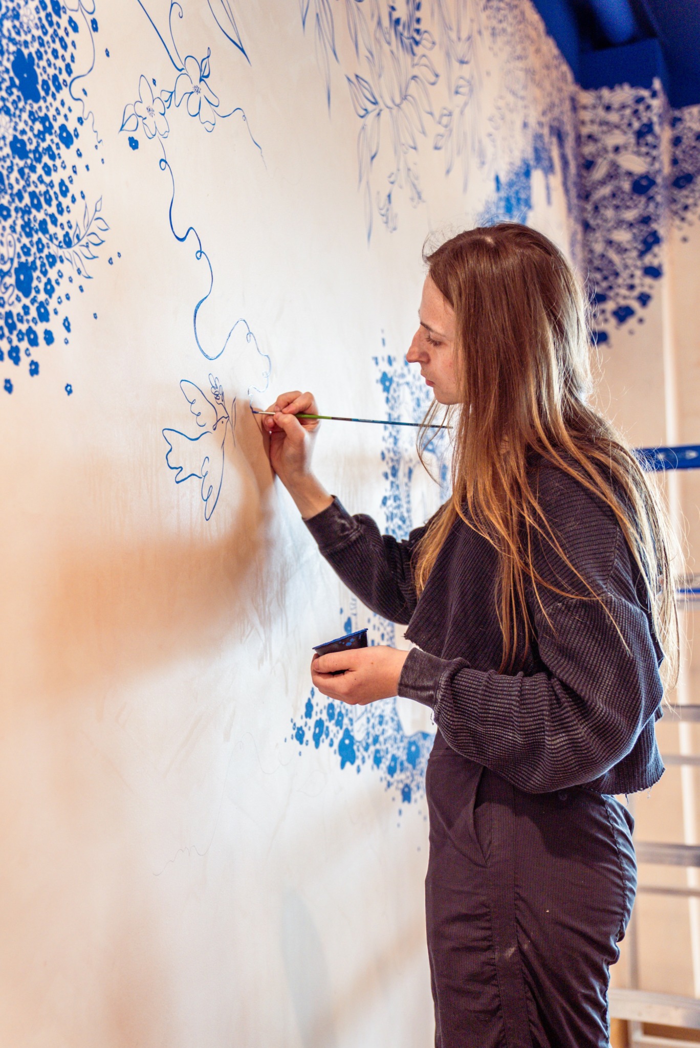 Young woman with long hair painting on a wall with blue paint, holding a brush and a container.