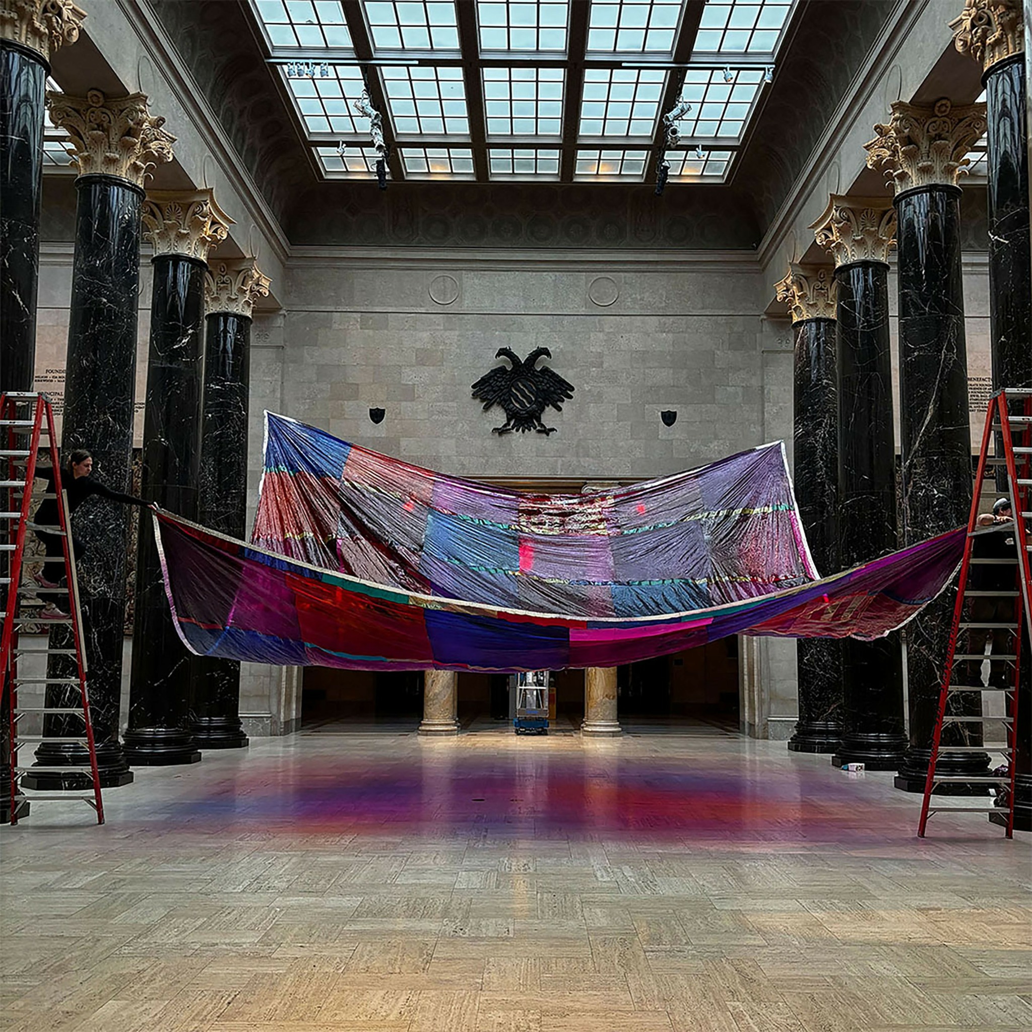 Large colorful fabric suspended between two red ladders inside a grand hall with tall black columns and a glass ceiling.