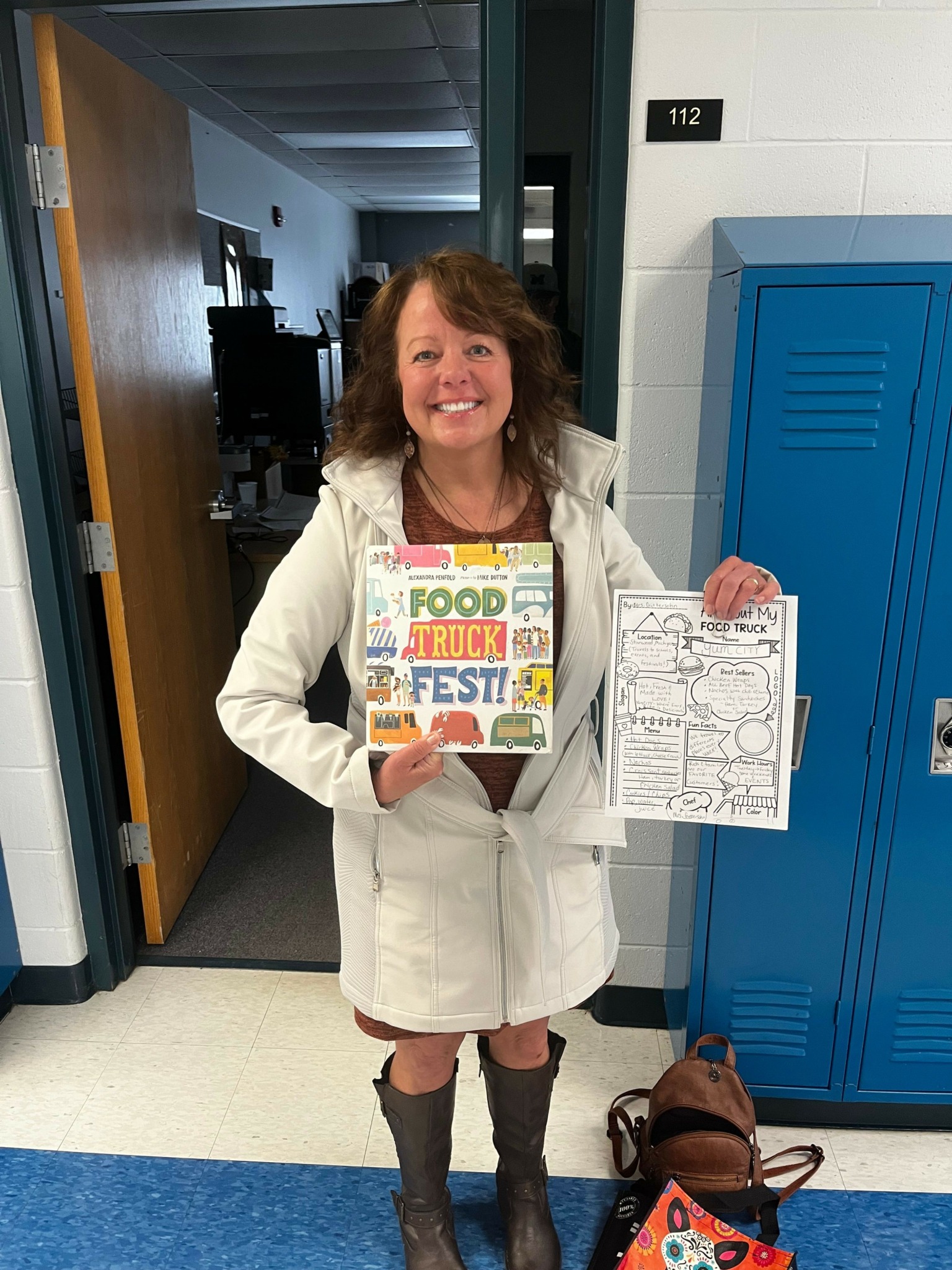 Woman holding a colorful book and a paper with drawings, standing in a hallway with blue lockers and a wooden door.