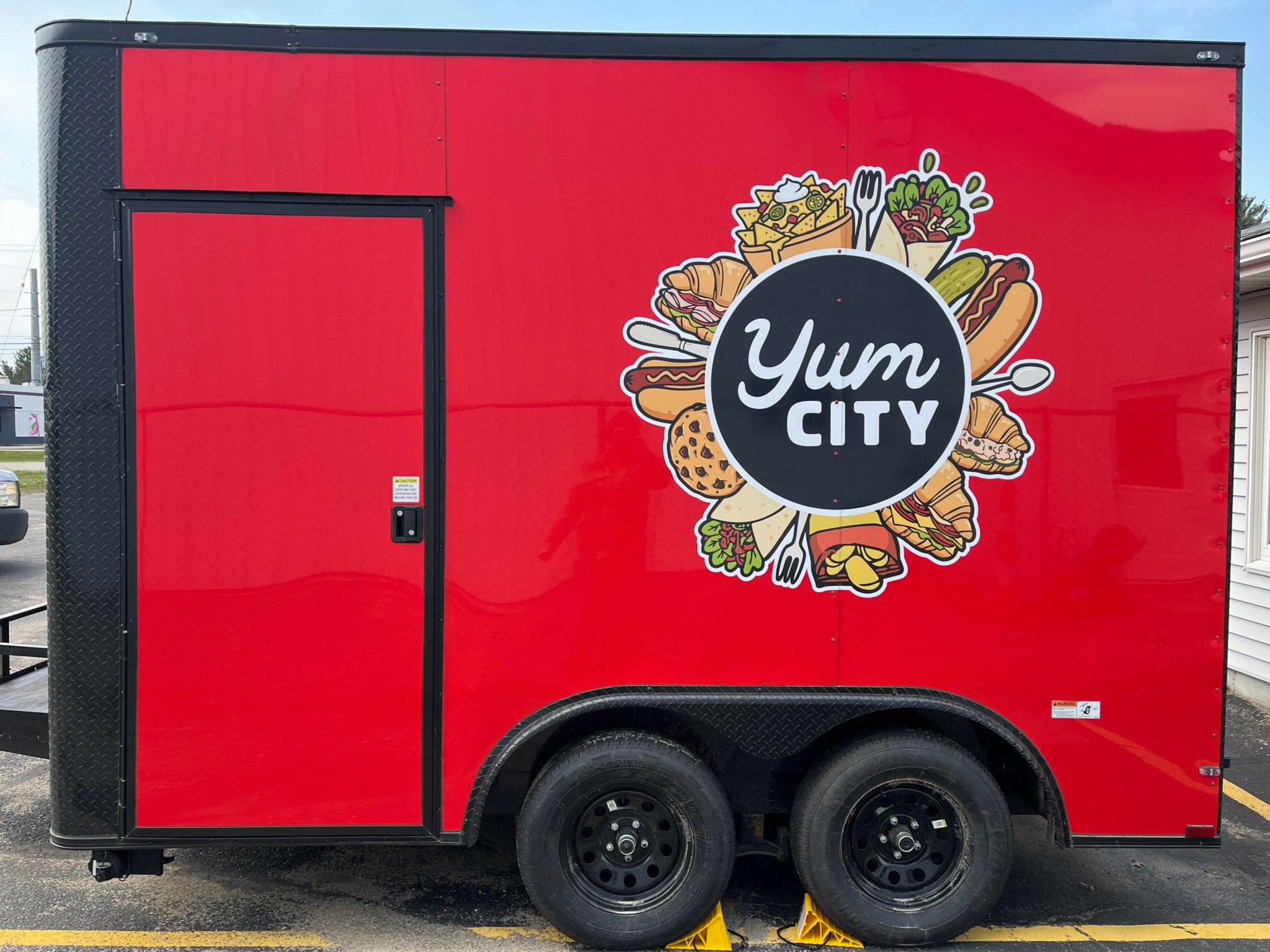 Red food truck with Yum City logo featuring various food illustrations, black wheel, and a door on the left side.
