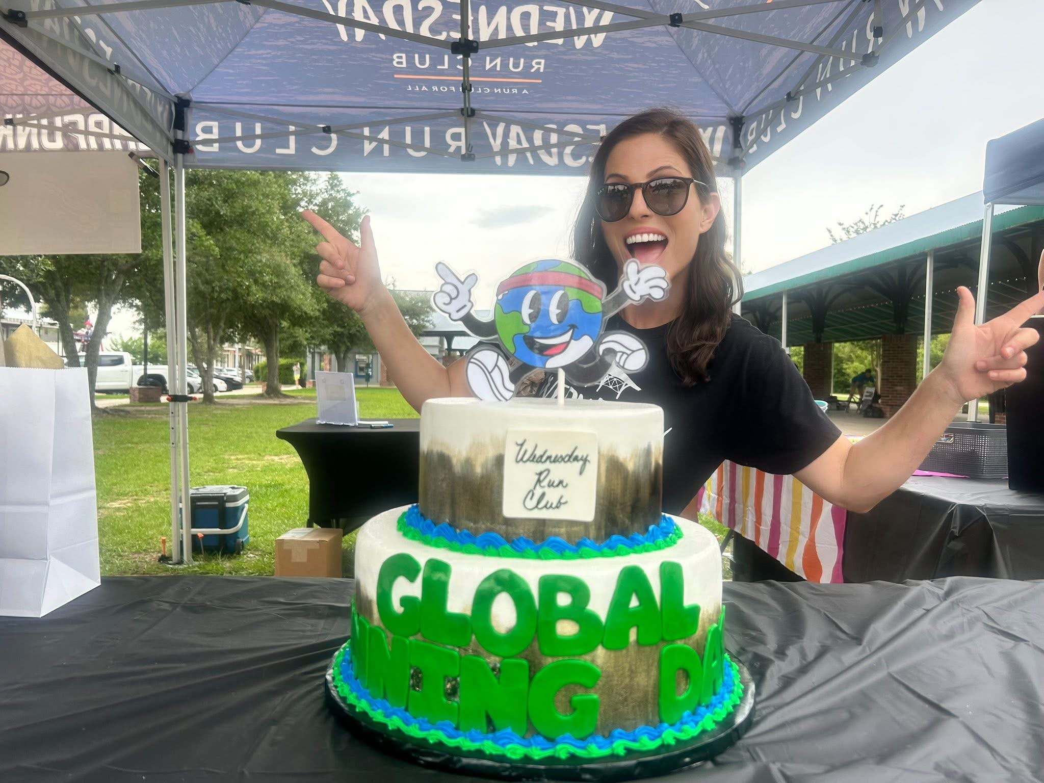 Woman celebrating outdoors with a two-tiered cake, wearing sunglasses, smiling, and pointing, under a canopy with trees in background.