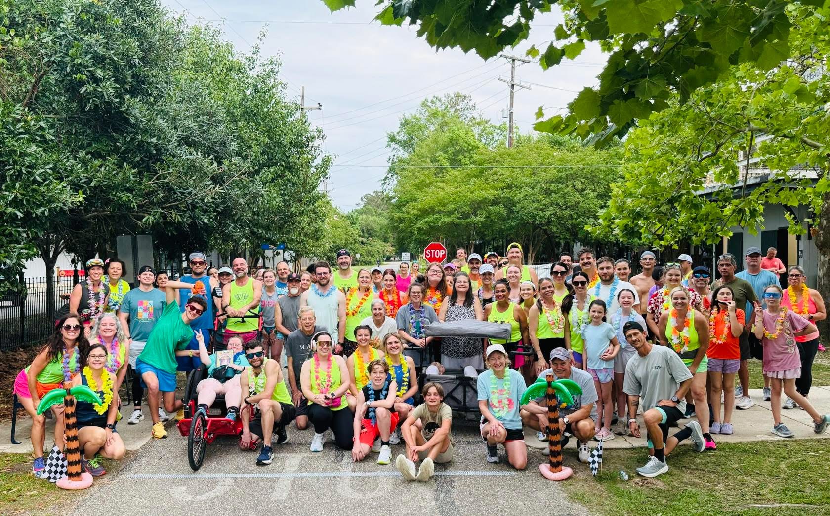Large group of people gathered outdoors on a street with trees, some holding umbrellas, posing for a photo.