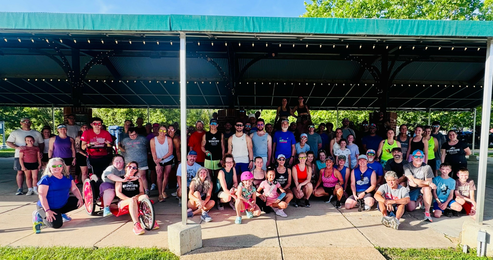 Group of people, including children, gathered under a large outdoor shelter on a sunny day.