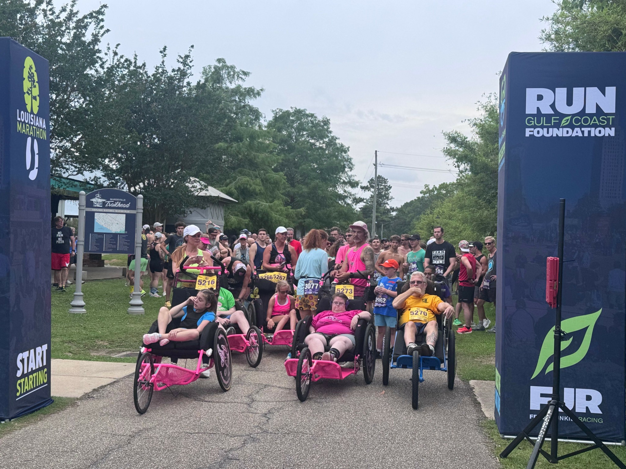 Group of people with strollers and wheelchairs gathered outdoors near race start, trees and signs visible in background.