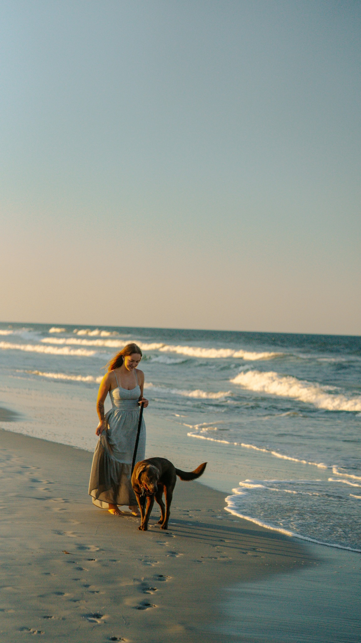 Woman walking with a dog on the beach during sunset, waves in the background, clear sky, footprints in sand.