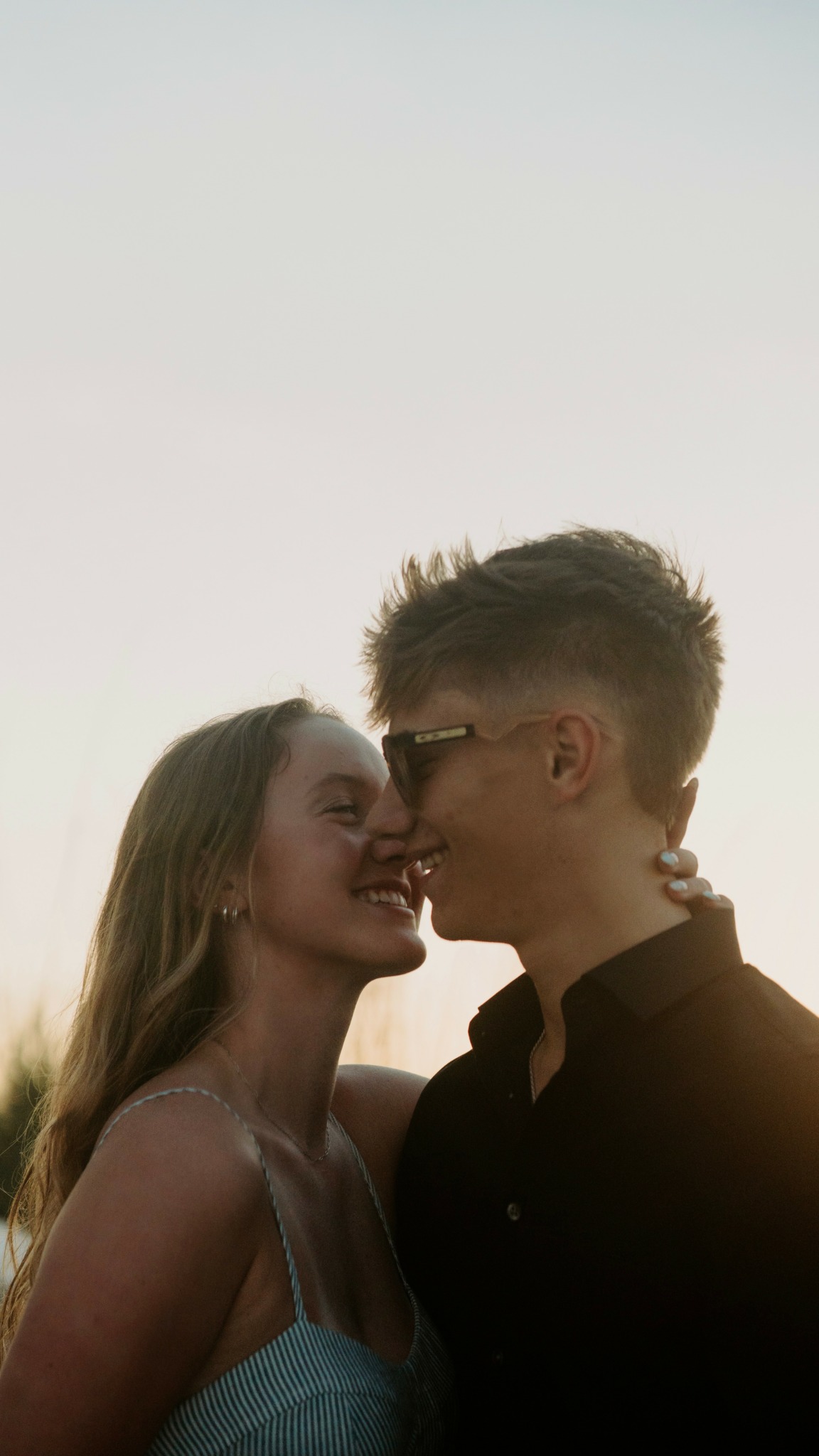 Two people face each other closely outdoors, smiling, with a clear sky background.