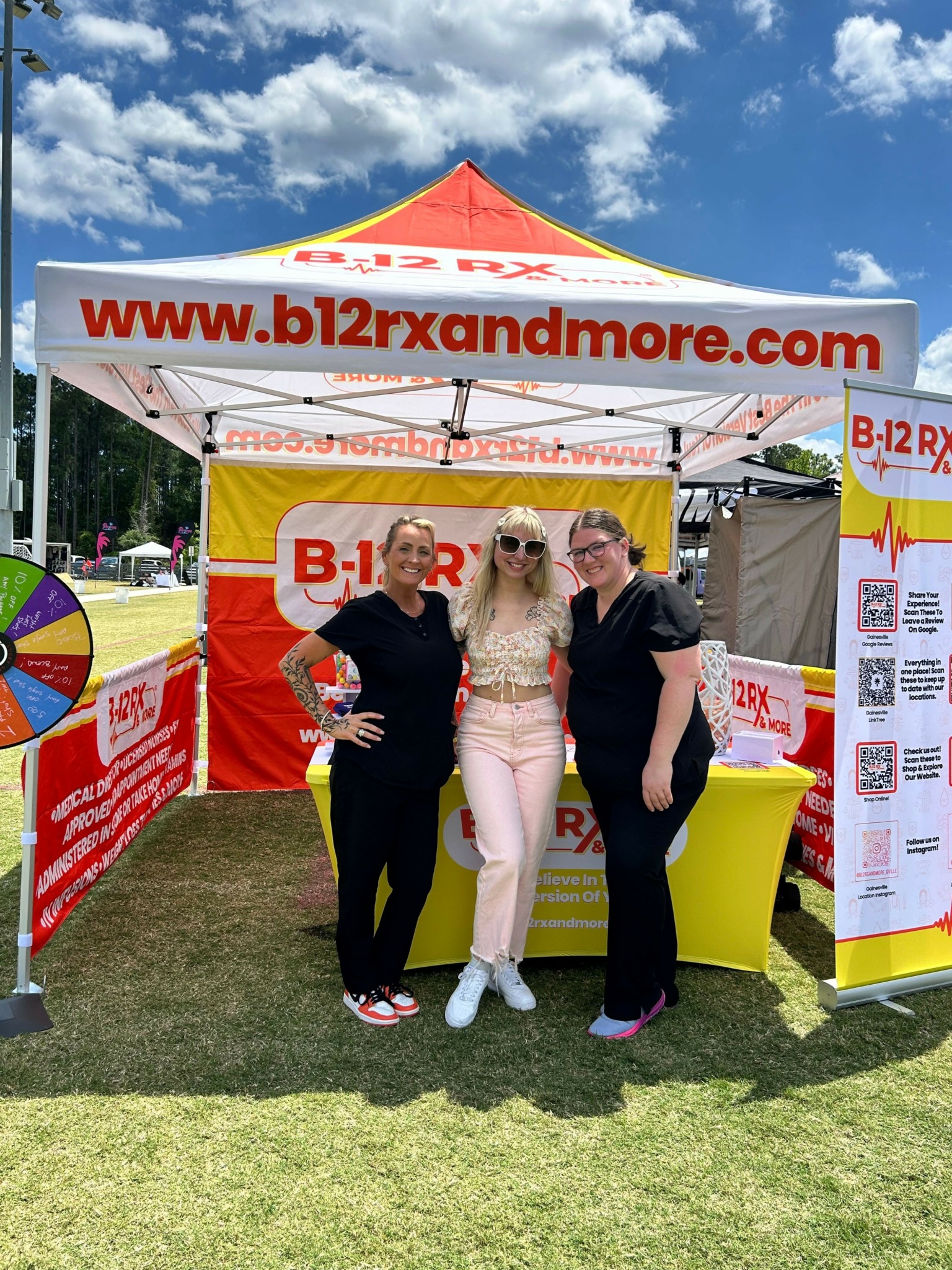 Two women stand in front of a promotional tent with banners, one holding a brochure, on a grassy field under a blue sky.