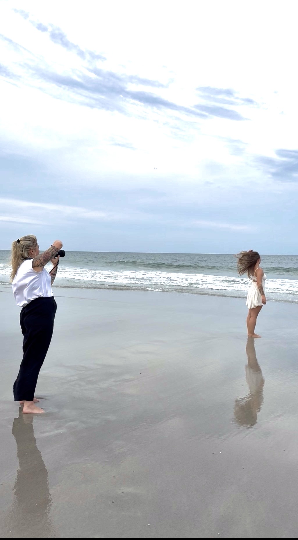 Two women on a beach, one taking a photo of the other standing in shallow water, with clouds and ocean in background.
