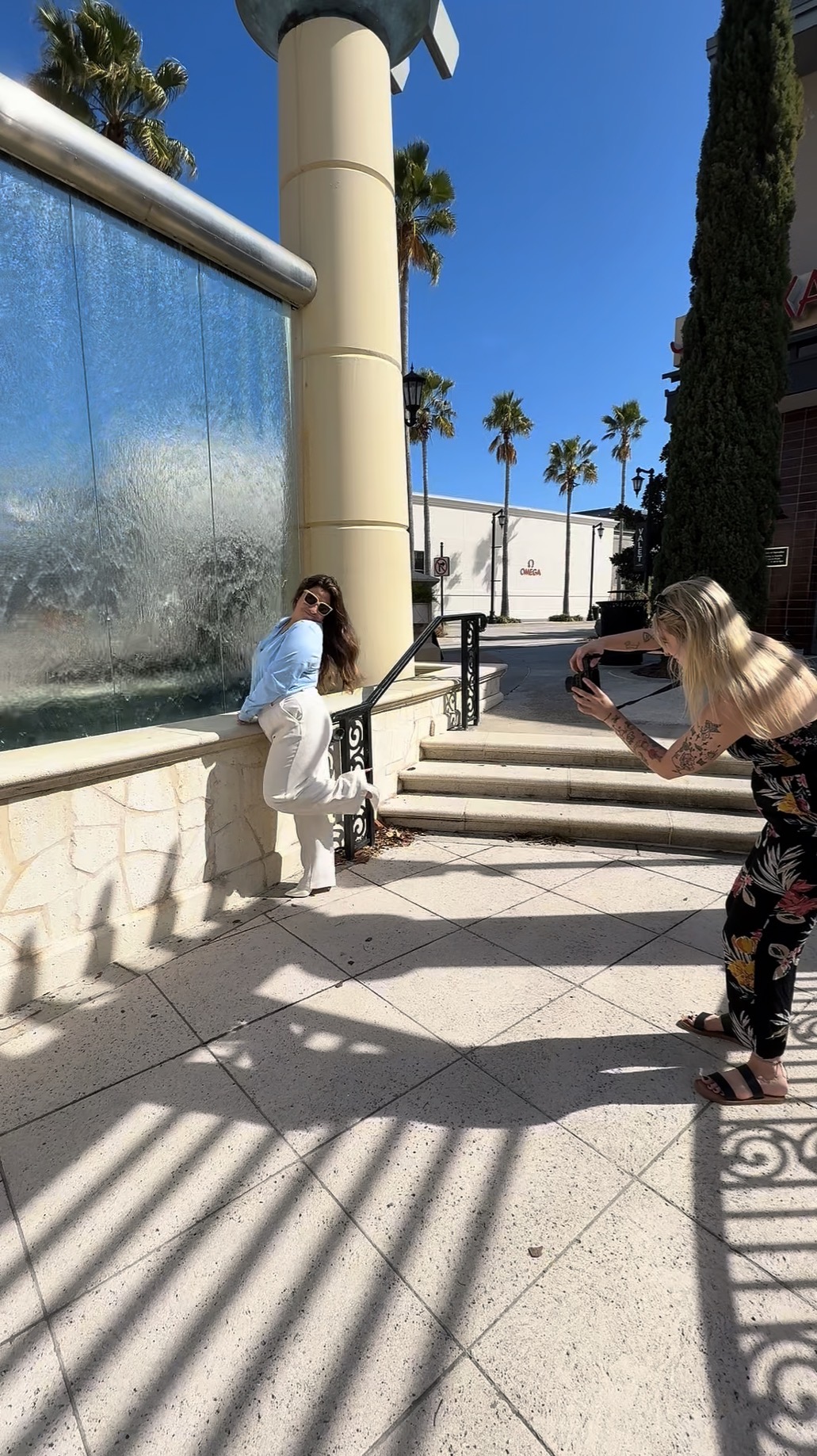 Two women on a sunny sidewalk, one taking a photo, with palm trees and a blue sky in the background.