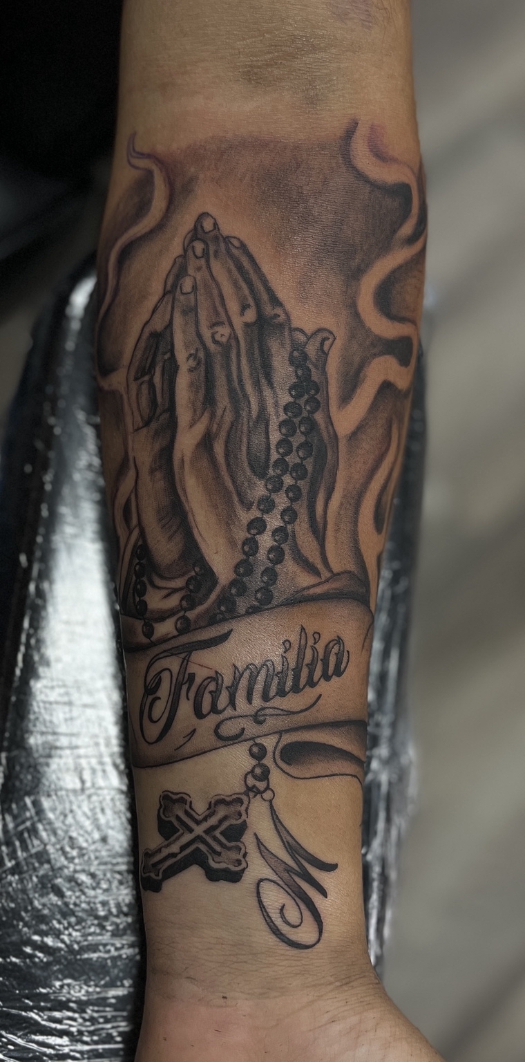 Tattoo of praying hands holding a rosary, with the word 'Familia' and decorative elements, on a person's forearm.