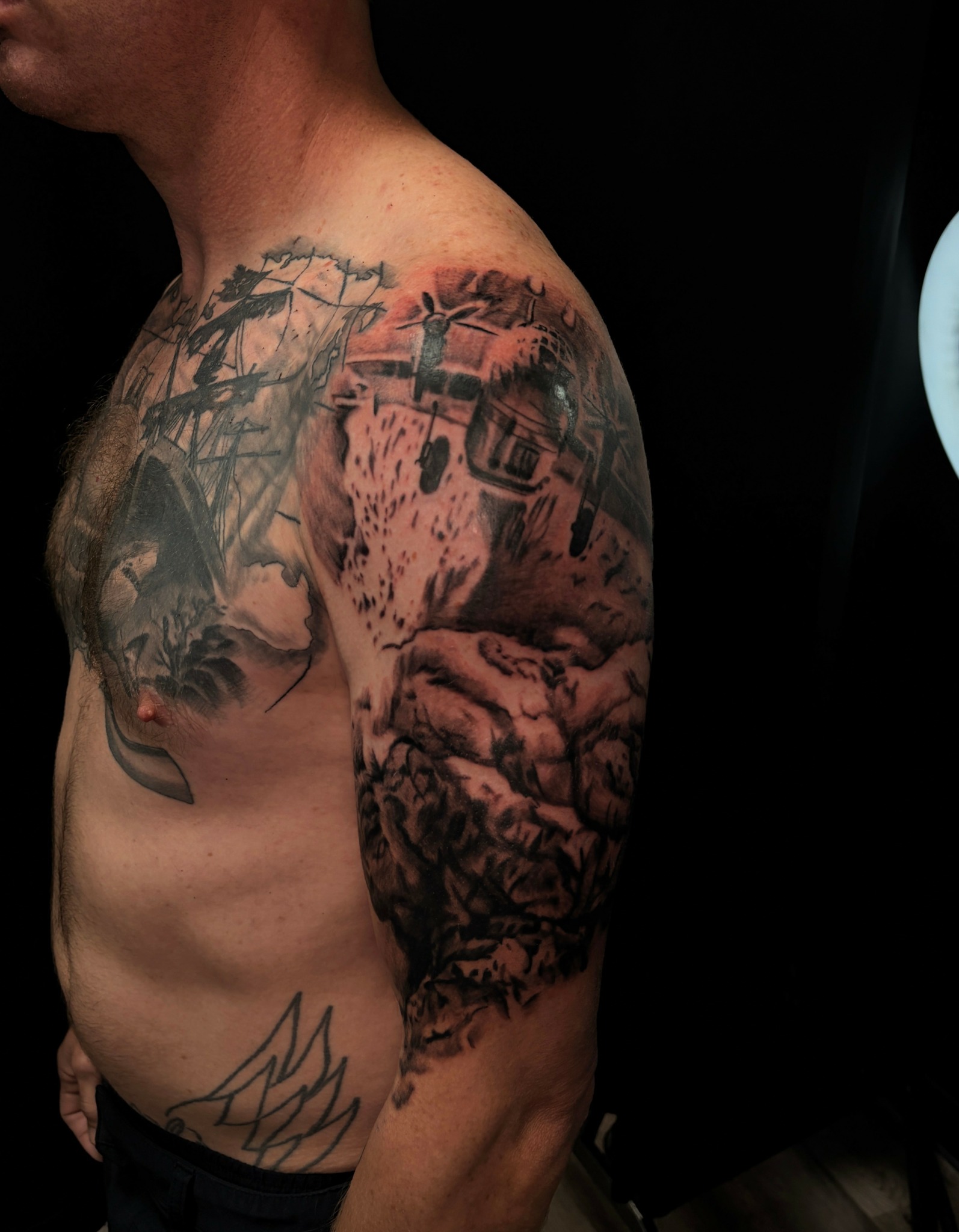 Tattooed person's upper arm and shoulder with detailed landscape and mountain designs, partially visible chest, and a small abstract design.