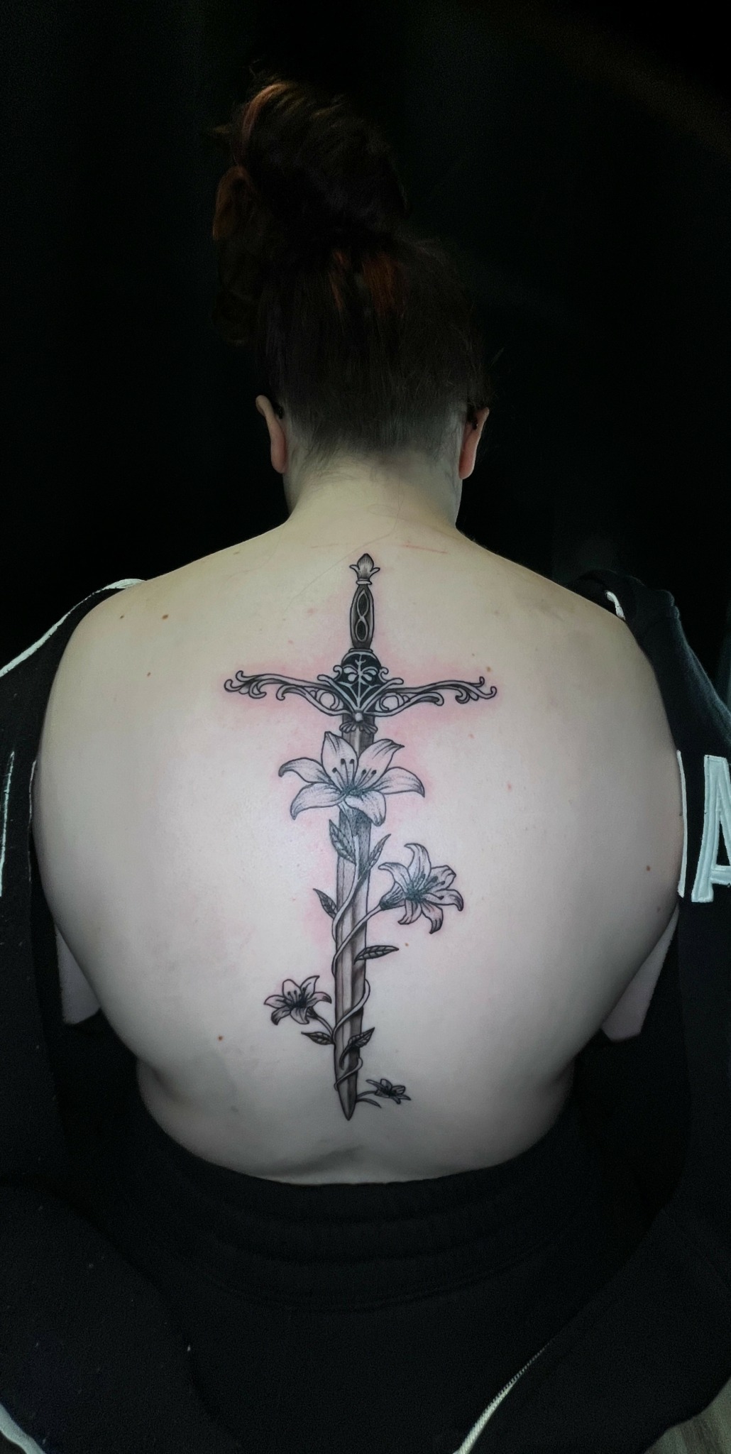 Back view of a person with a large, detailed tattoo of a sword with floral elements on their back.