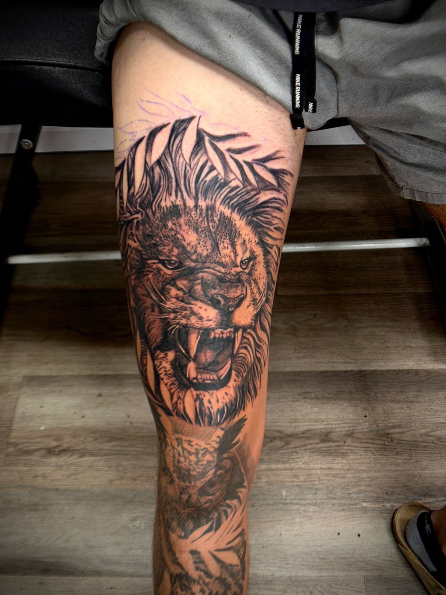 Tattoo of a roaring lion's face with detailed mane on person's thigh, with leaves around it.