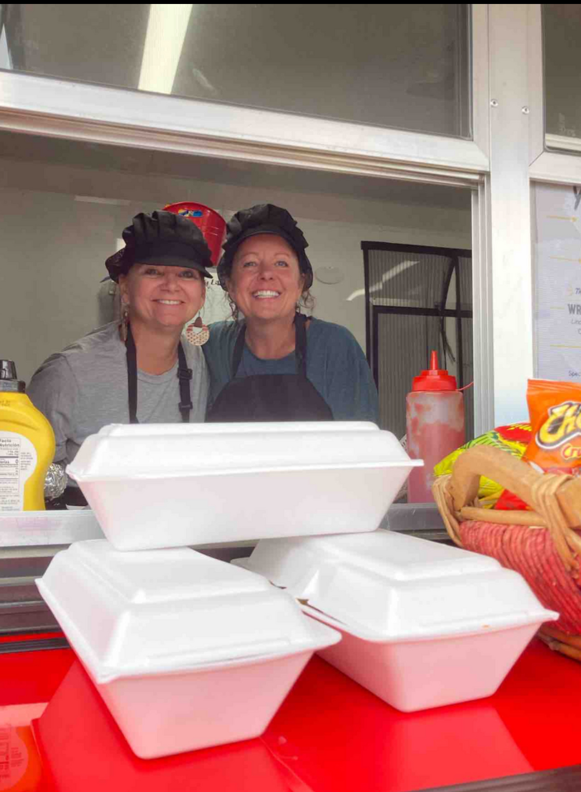 Two women smiling behind a food counter with takeout containers and condiments, viewed through a window.