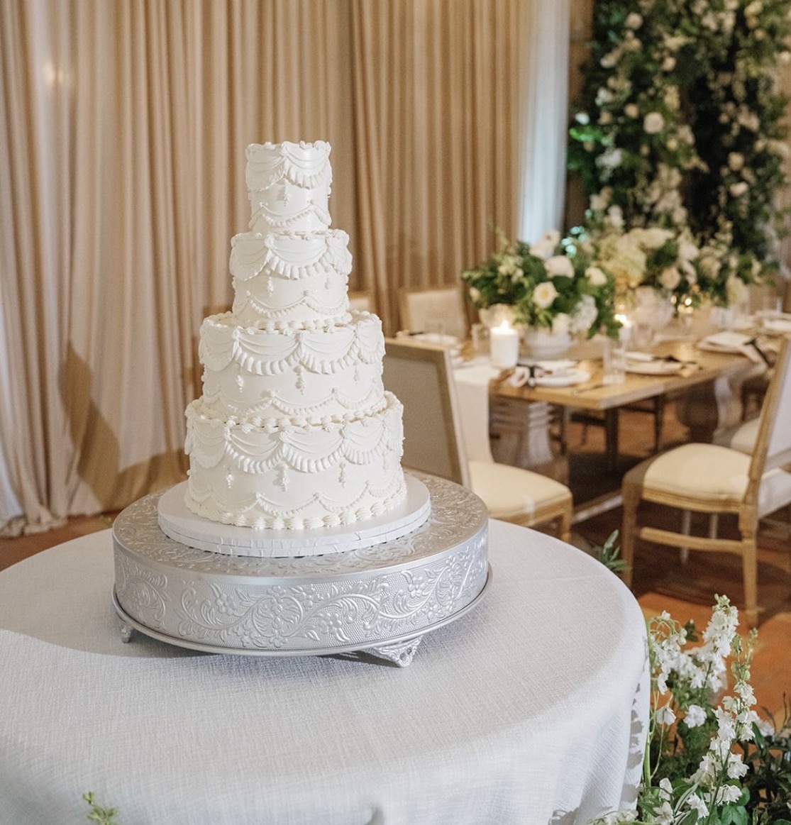Four-tiered white wedding cake with decorative icing, on a silver stand, in a decorated event space with floral arrangements.