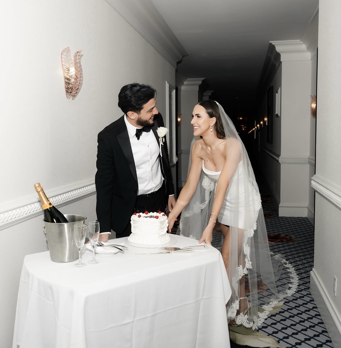 Bride and groom cut wedding cake at a table, champagne bottle in ice bucket, elegant hallway background.