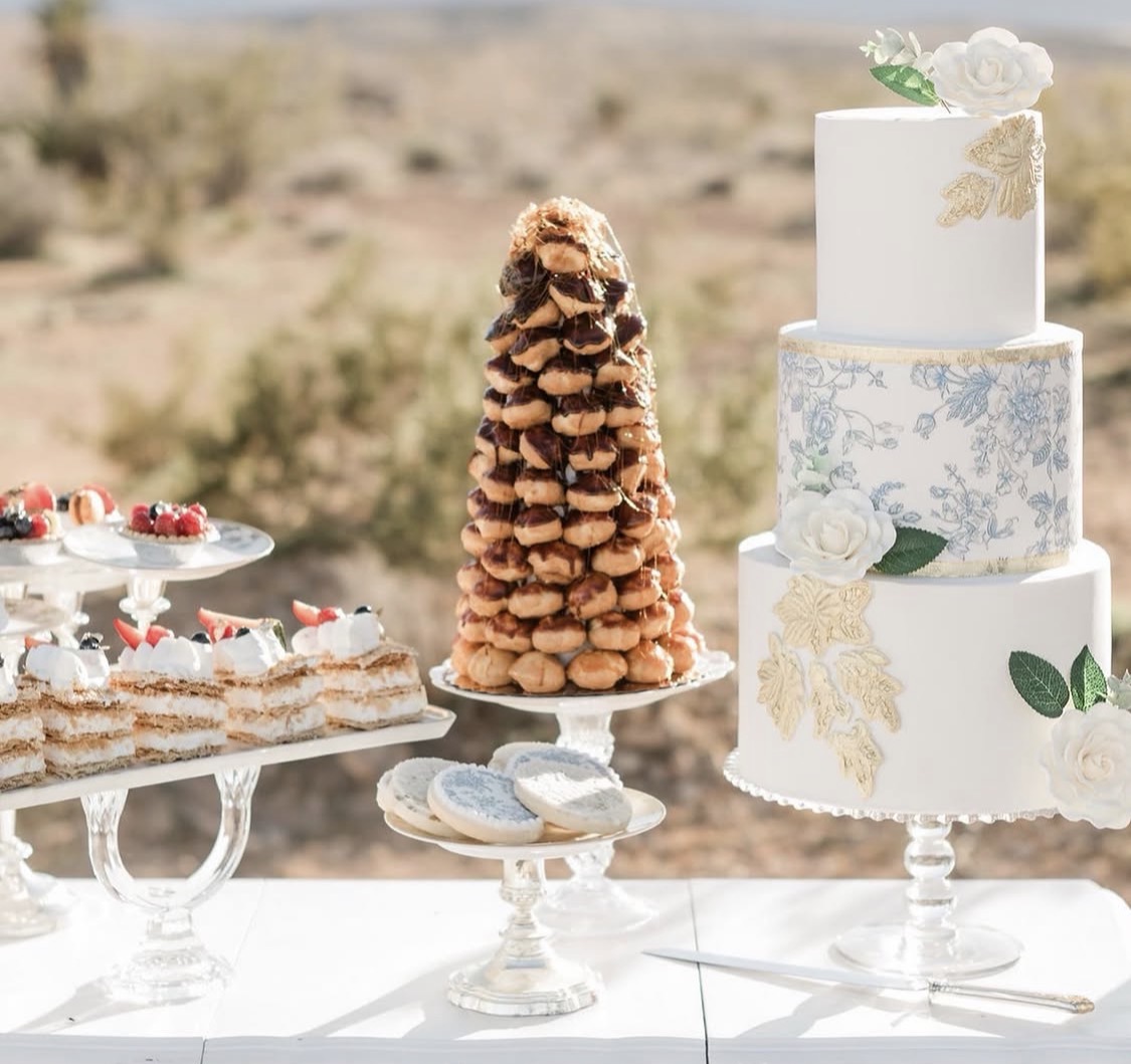 Dessert table with a tall layered cake, cookies, and pastries outdoors, with a blurred natural background.