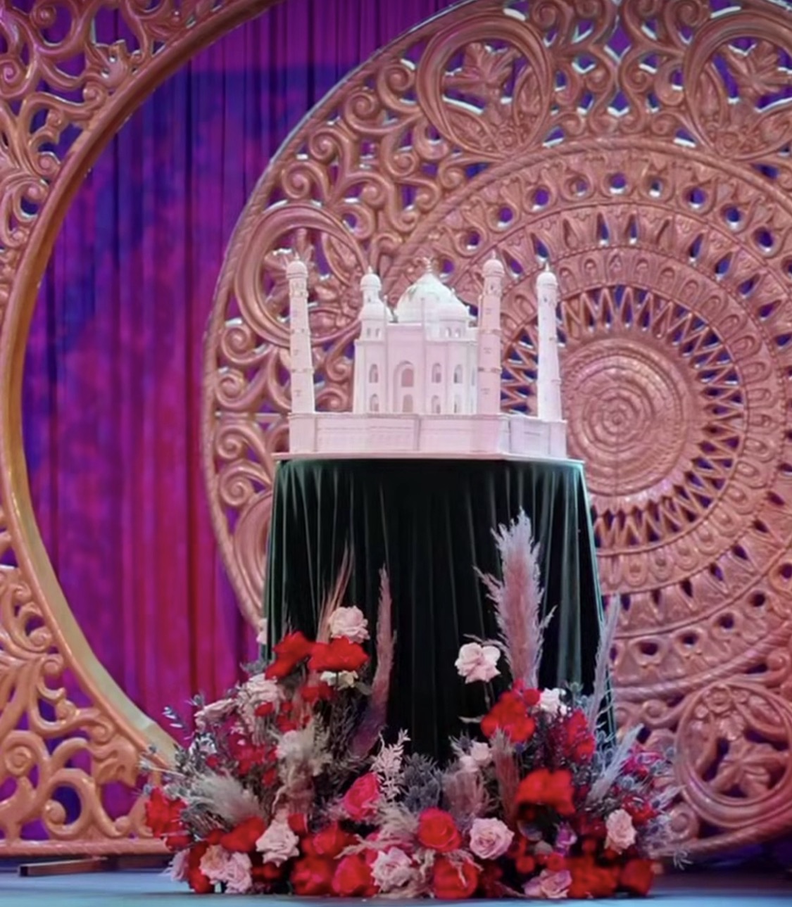 A white cake with pink decorations on a black table, surrounded by red and pink flowers, with a decorative backdrop.