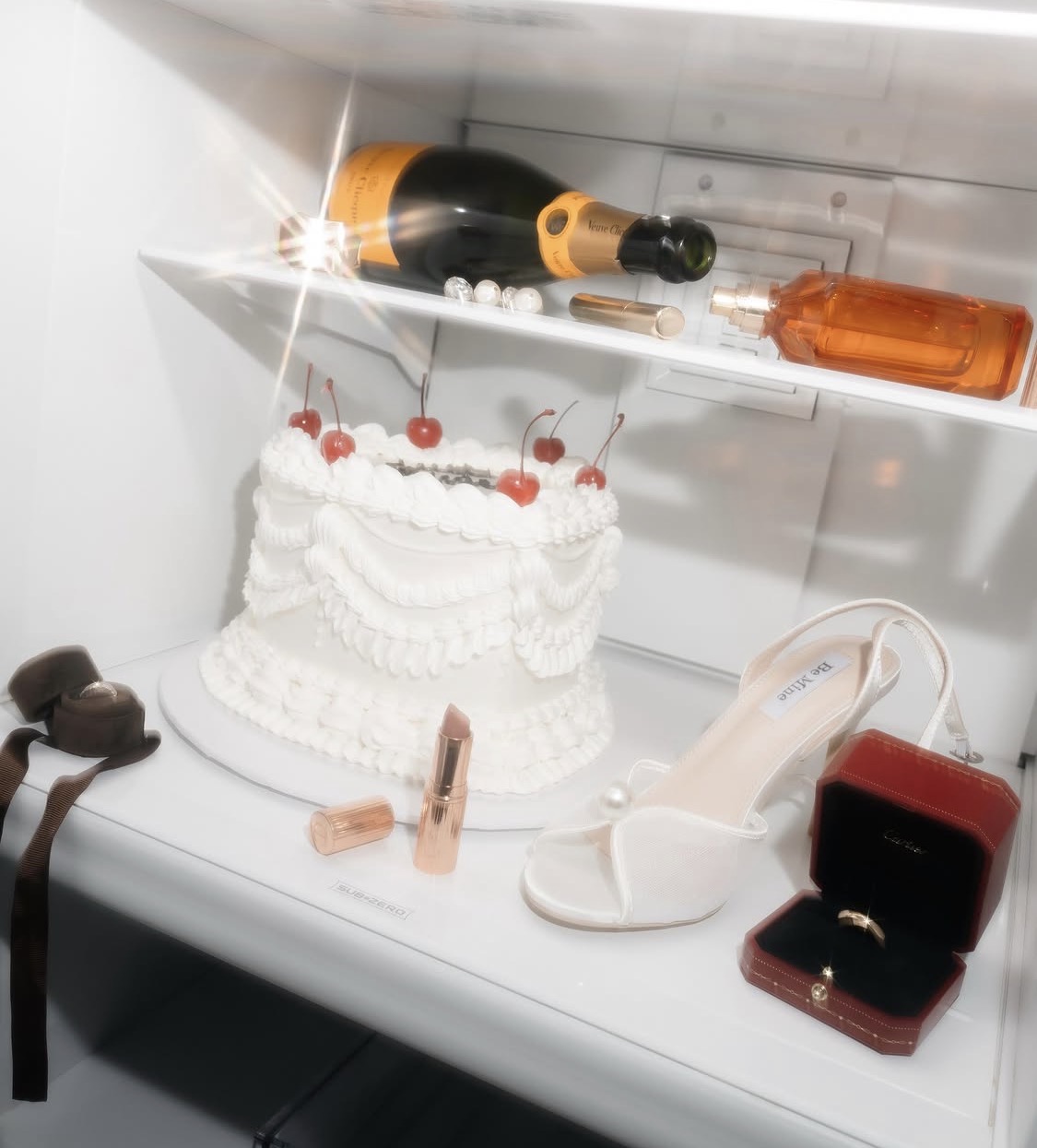 A white cake with red cherries on top, a high-heeled shoe, a bottle of champagne, and jewelry inside a refrigerator.