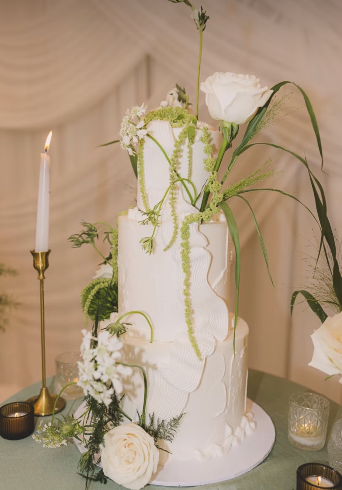 Tall white wedding cake decorated with flowers and greenery, with a lit candle nearby, on a table.