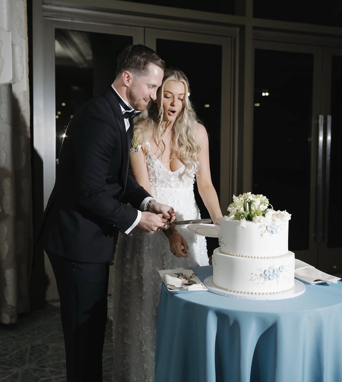 A bride and groom cut a wedding cake together indoors at night, with a table covered in a blue cloth.
