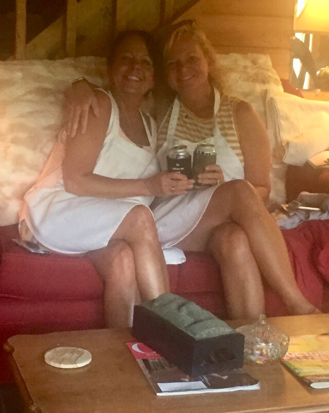 Two women sitting on a couch, smiling, holding drinks, with a wooden wall and lamp in the background.
