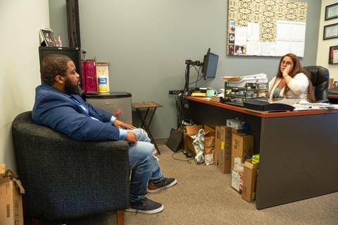 Man and woman sitting in an office, facing each other, with desk and office supplies nearby.