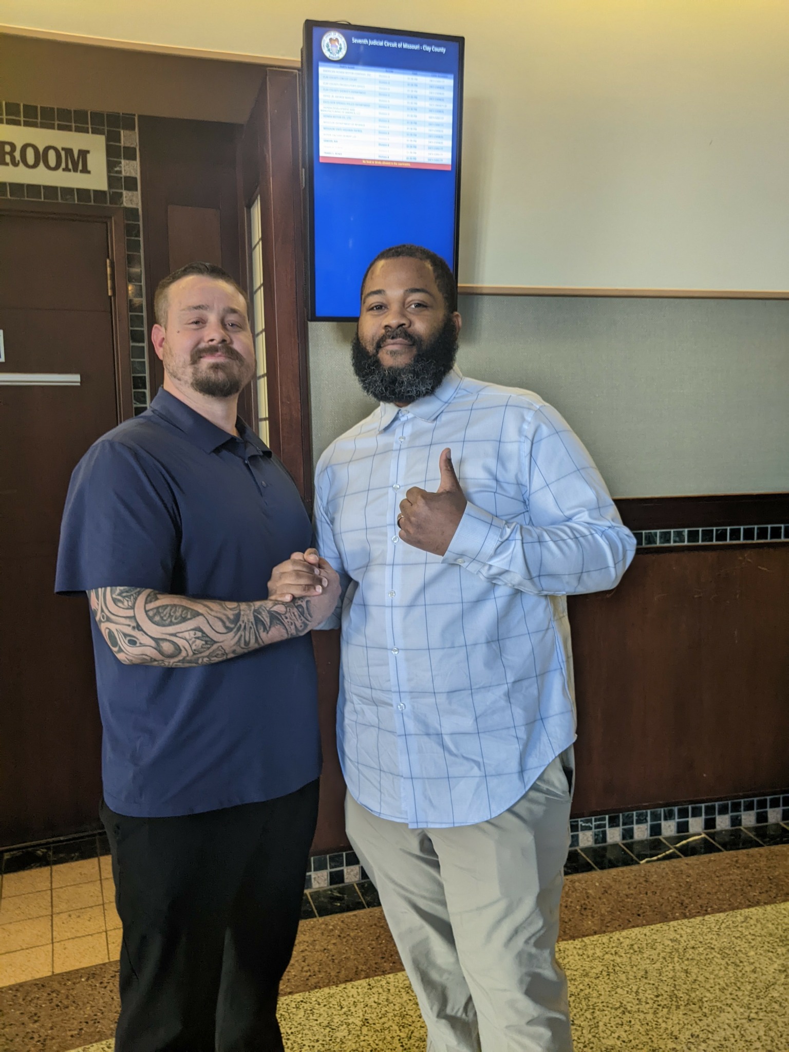 Two men shaking hands indoors, one with a tattooed arm, the other with a beard, standing in front of a digital display.