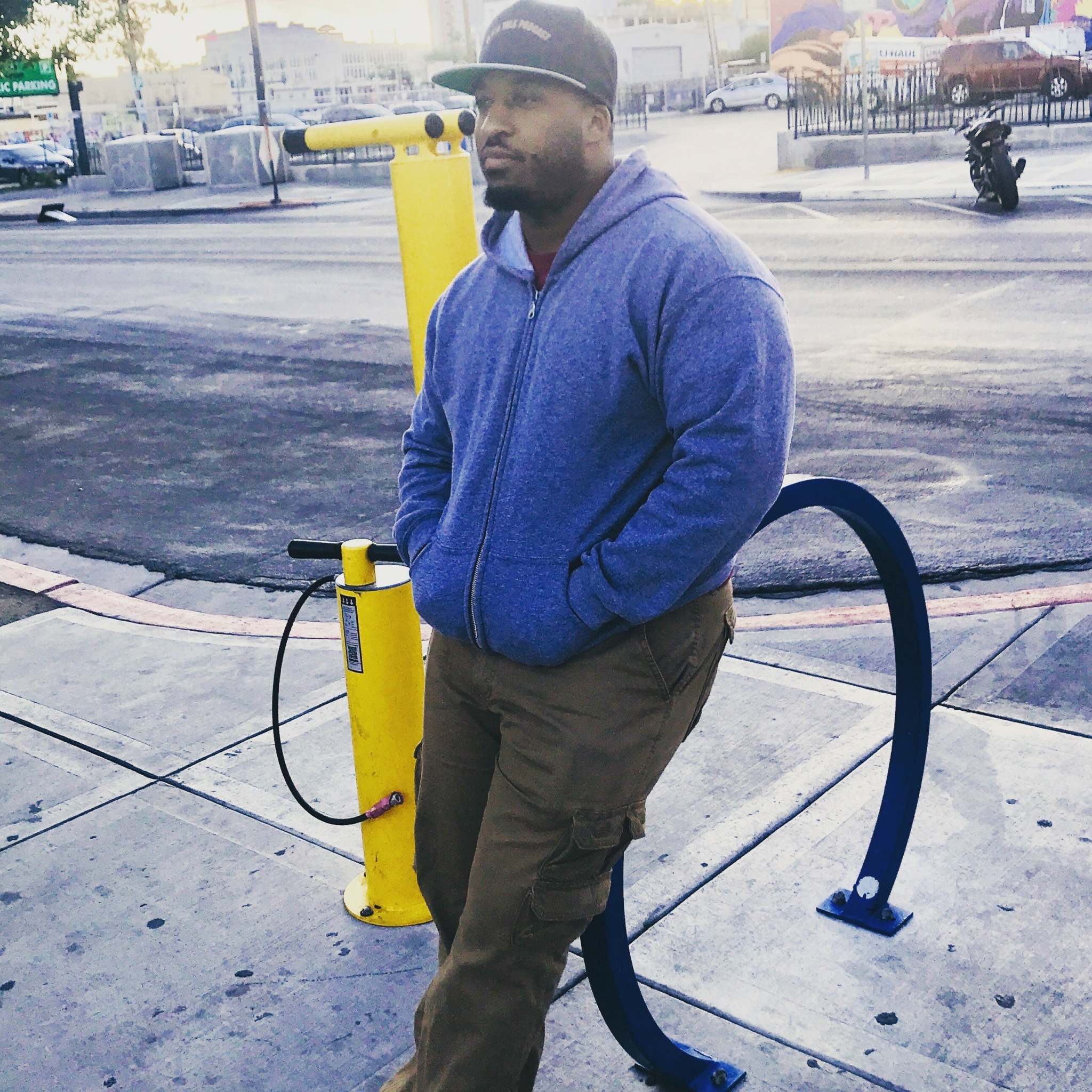 Man in blue hoodie and brown pants standing outdoors near a yellow bike rack, hands in pockets, looking to the side.