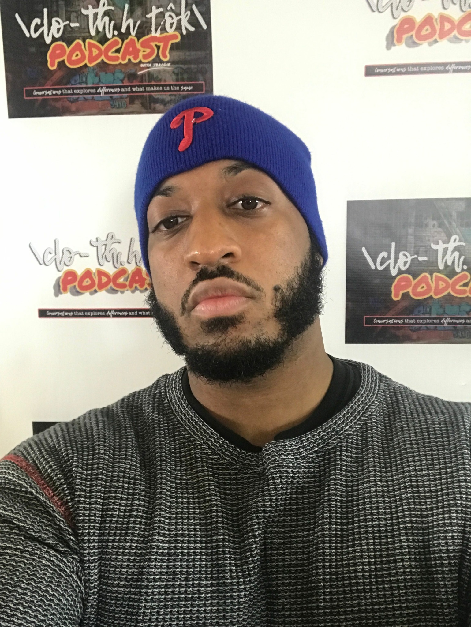 Young man with a beard wearing a blue beanie and a black and gray shirt, standing in front of posters on a wall.