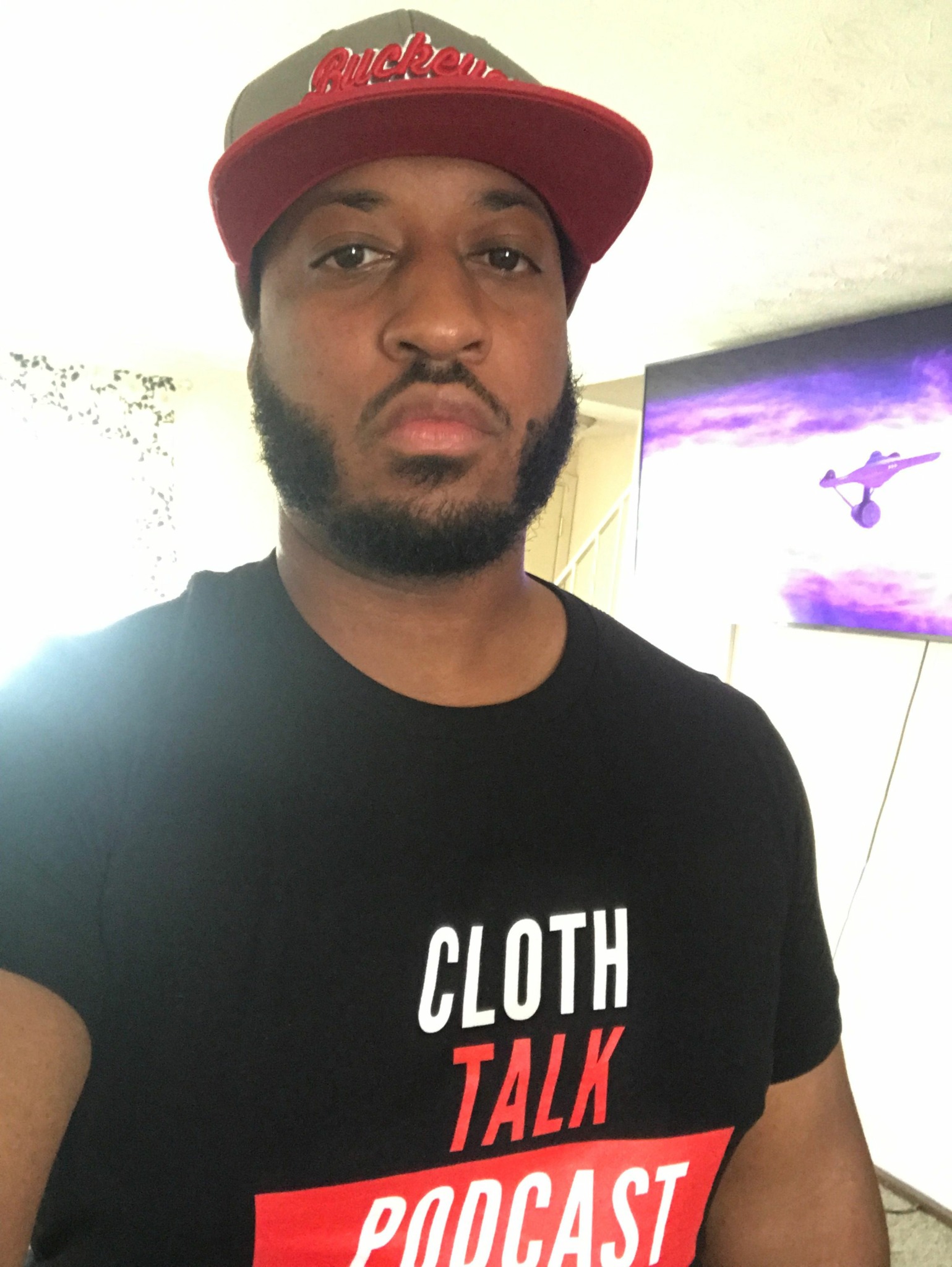 Person wearing a red cap and black T-shirt with white and red text, standing indoors with a TV in the background.