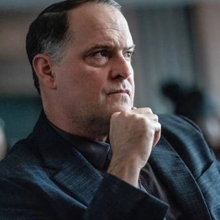 Man with short dark hair, wearing a dark suit, resting his chin on his hand, looking serious.