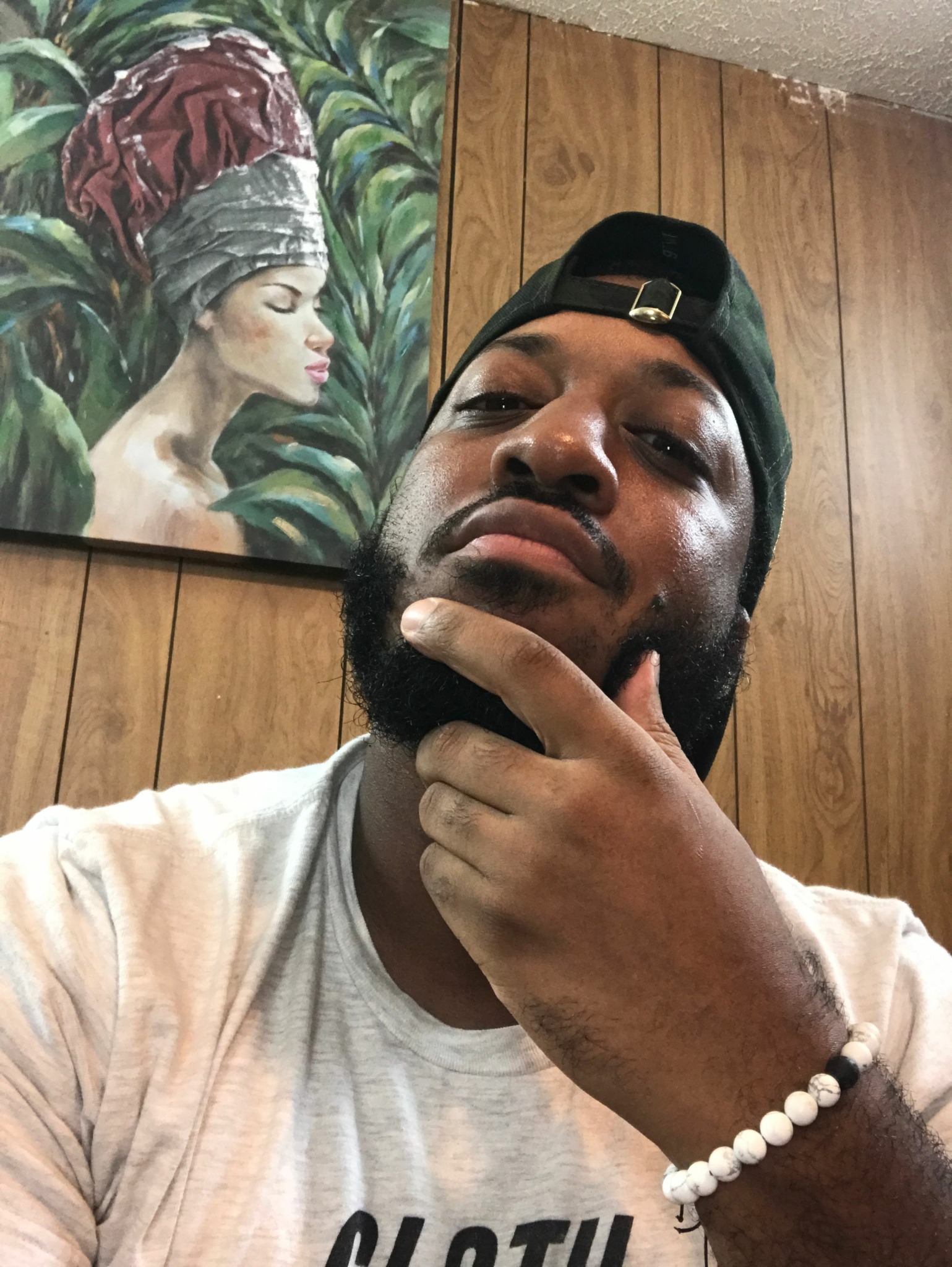 Young man with a beard and cap, lying on wooden floor, touching his chin, with a painting of a woman in the background.