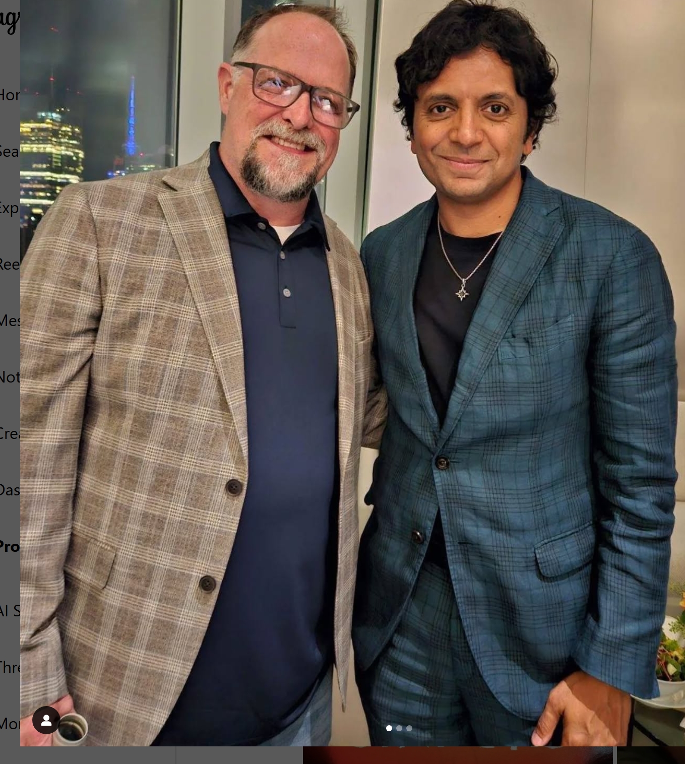 Two men stand side by side indoors, smiling at the camera, with city lights visible through a window behind them.