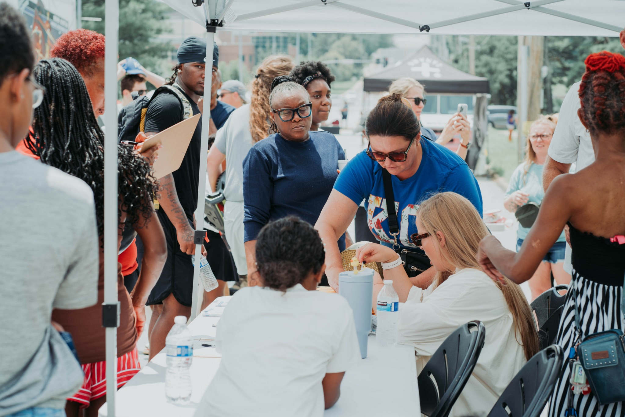 Group of people gathered around a table outdoors, some interacting and others waiting, under a canopy.