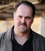 Man with dark hair, mustache, and beard, wearing a dark shirt and blazer, outdoors with blurred background.