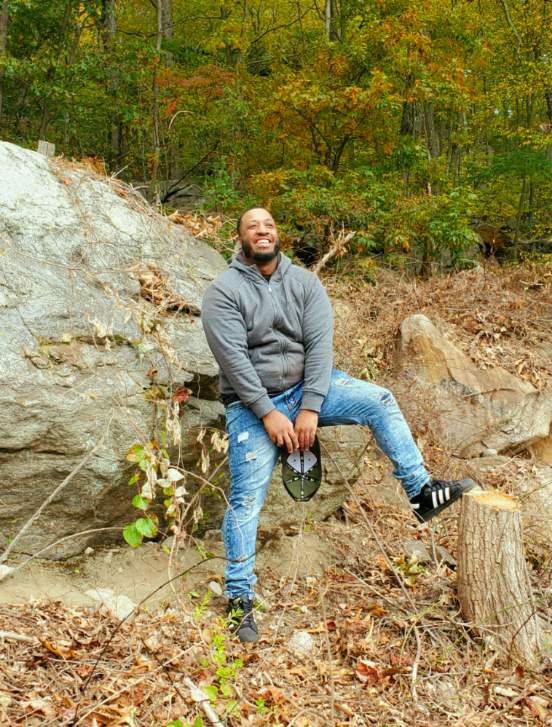 Person standing outdoors on rocky terrain with trees in the background, smiling and holding a hat, wearing a gray jacket and jeans.
