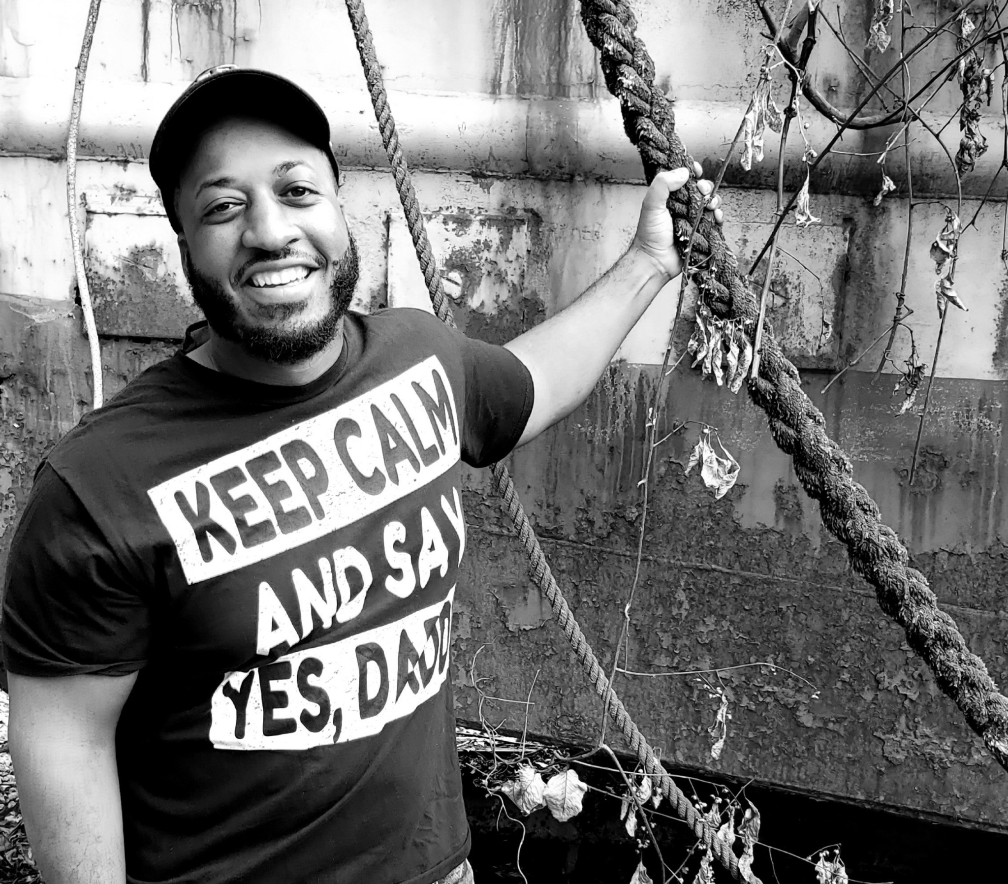 Smiling man with a beard wearing a cap and a T-shirt that says 'KEEP CALM AND SAY YES, DAD' holding a rope on a boat.