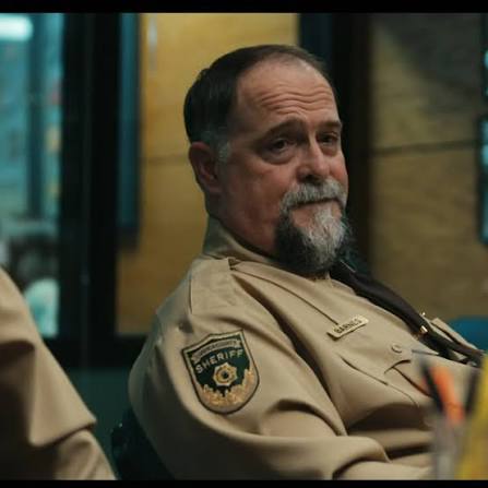 Man in sheriff uniform with a badge, sitting indoors, looking to the side, with a serious expression.