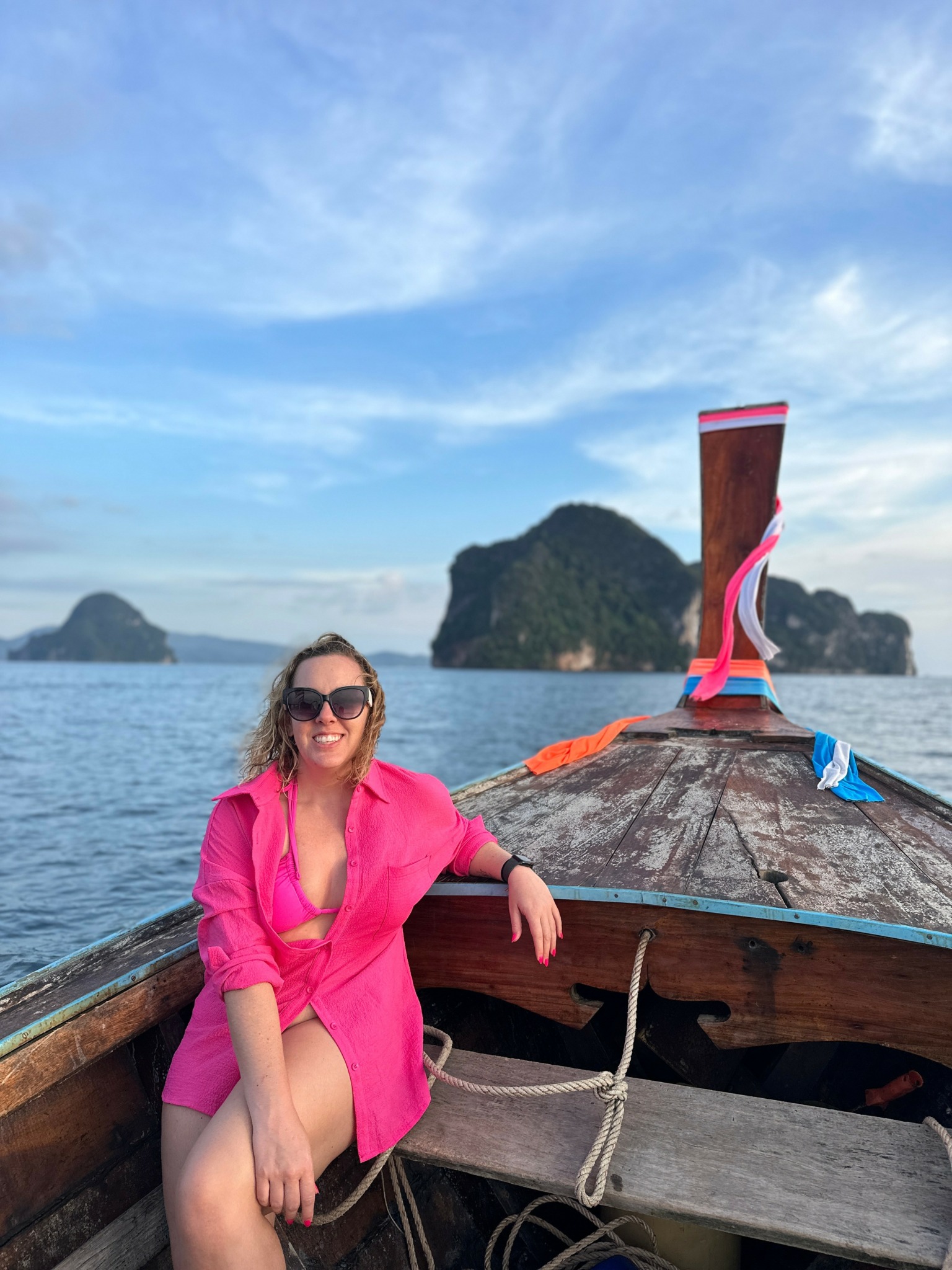 Woman in pink dress sitting on boat with ocean and islands in background, blue sky with clouds.