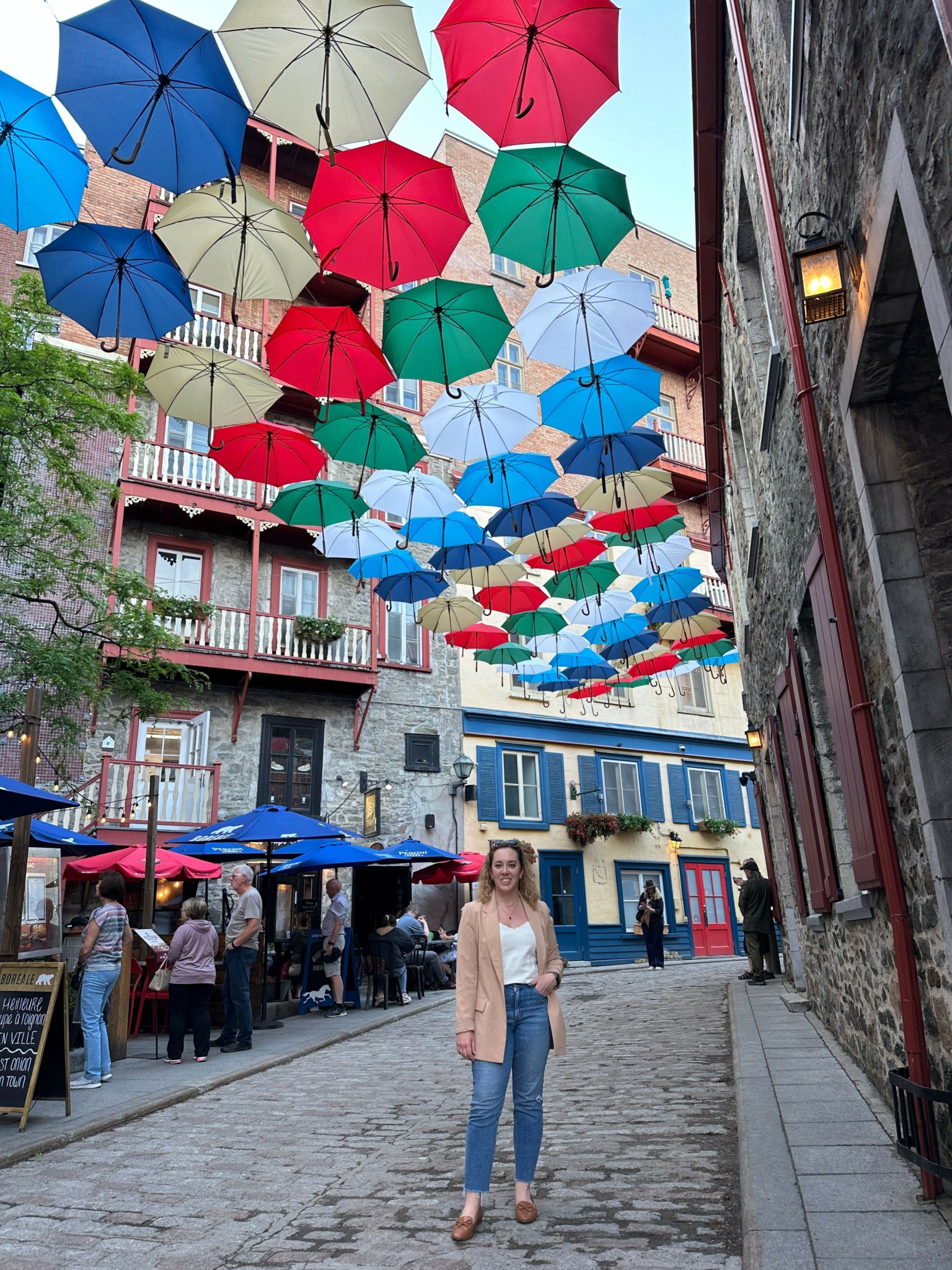 Person standing on cobblestone street with colorful umbrellas overhead, buildings in background, and outdoor seating area with blue umbrellas.
