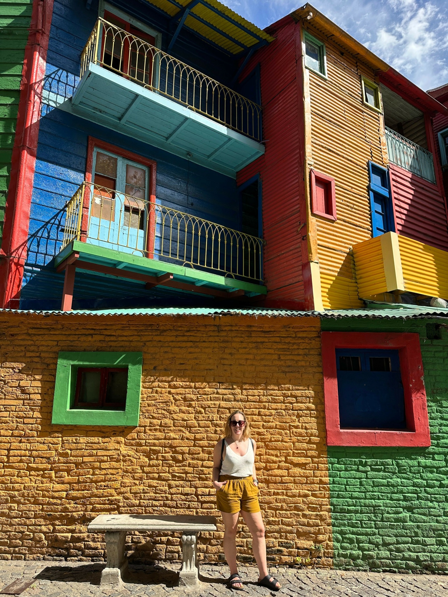 Child standing on a colorful brick wall with windows, next to a bench and a person, building with vibrant colors in background.