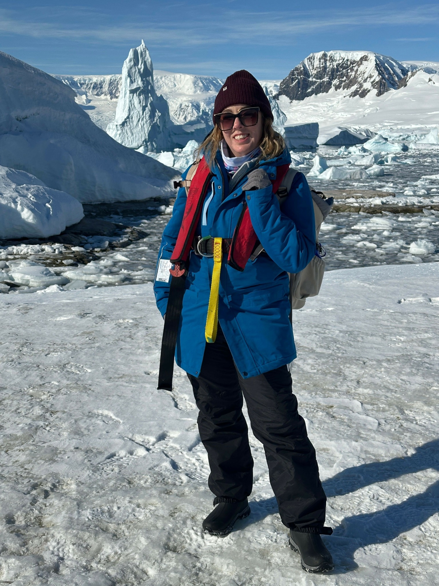Person standing on icy landscape with snow-covered mountains in background, wearing winter gear and smiling.