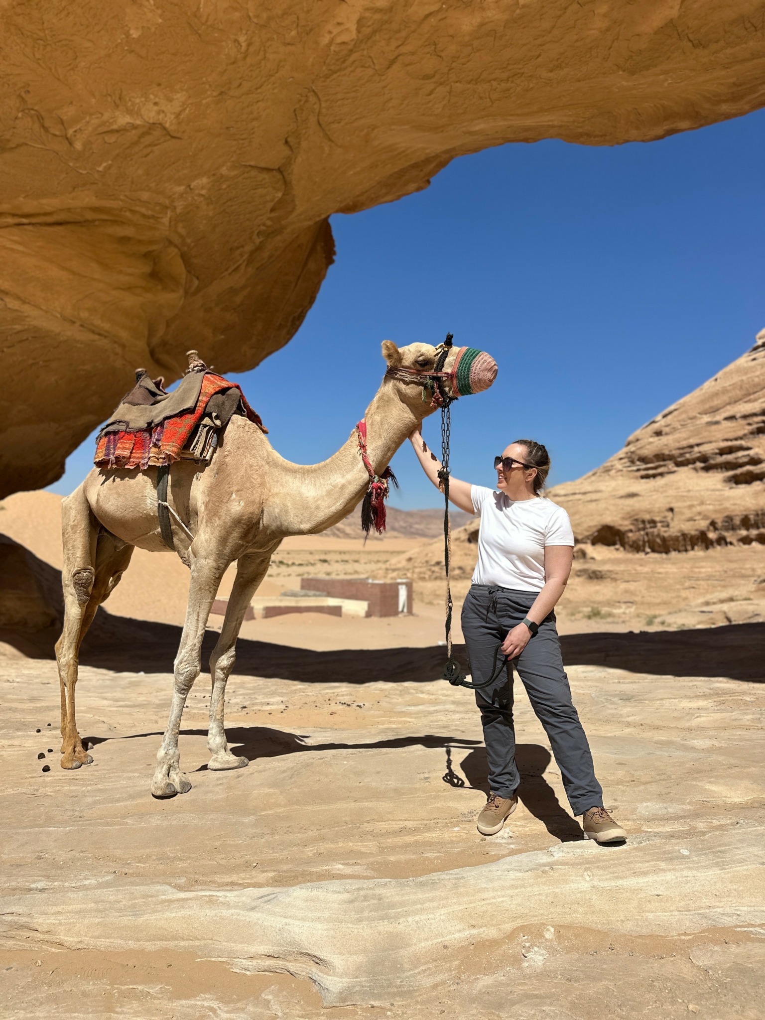 Person standing next to a camel under a large rock formation in a desert landscape, wearing sunglasses and casual clothing.