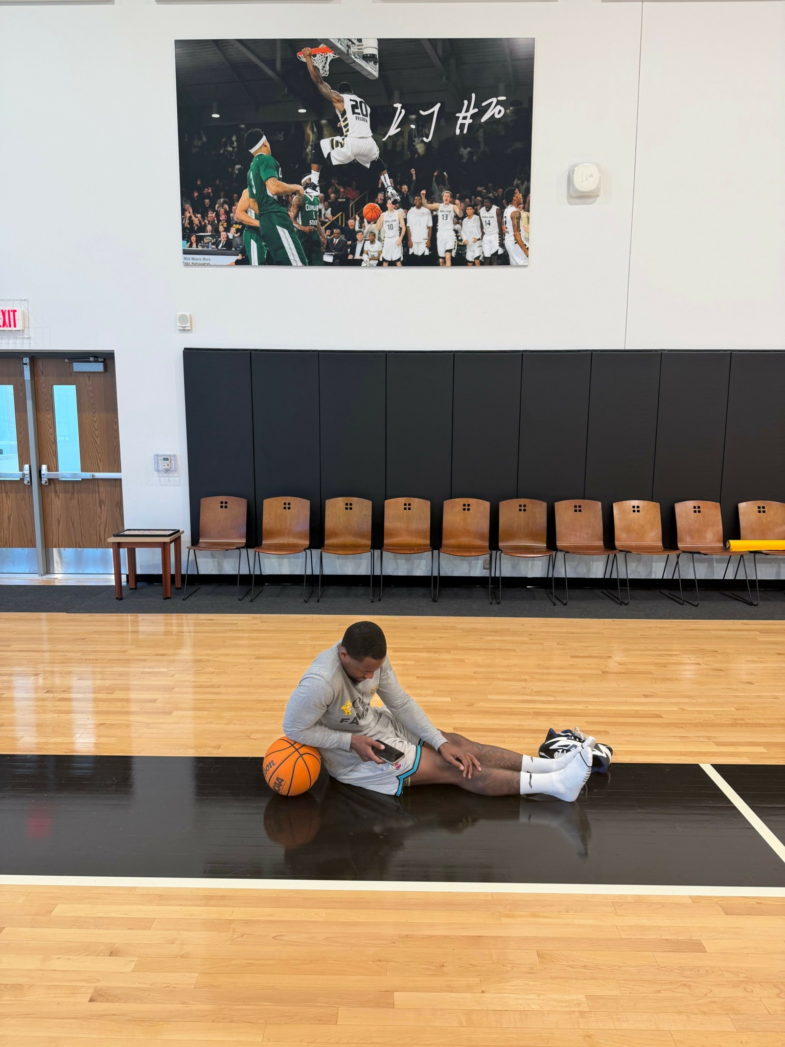 Person sitting on gym floor holding basketball, with gym equipment and a poster on the wall behind.