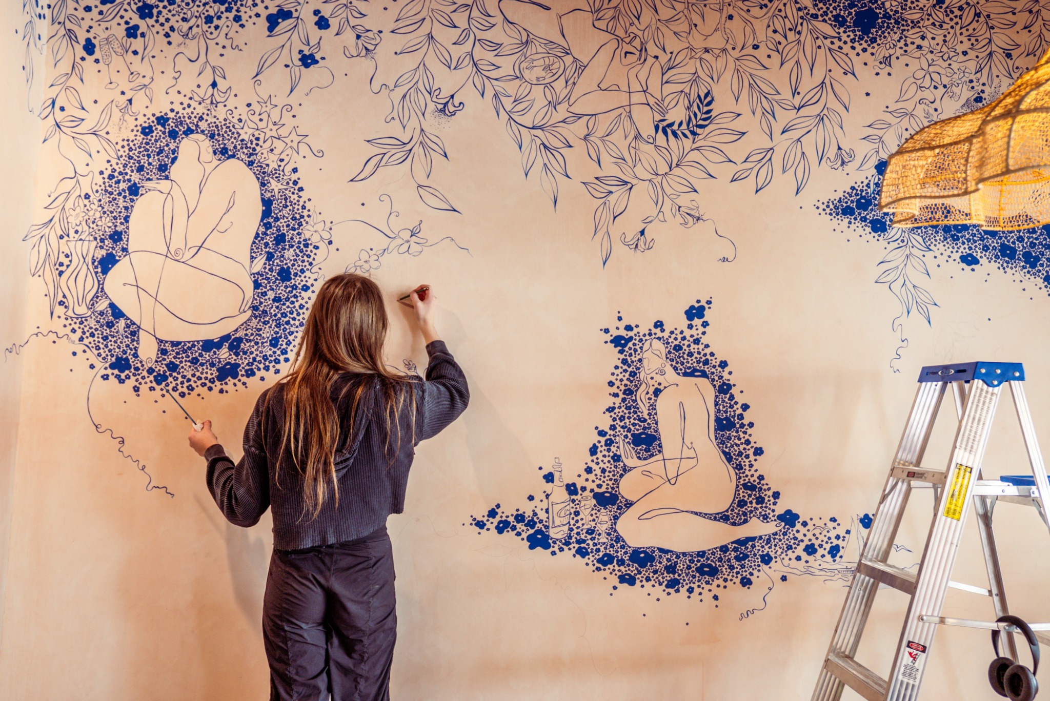 Woman painting a mural with blue floral and abstract designs on a wall, ladder nearby, and a yellow umbrella in the corner.