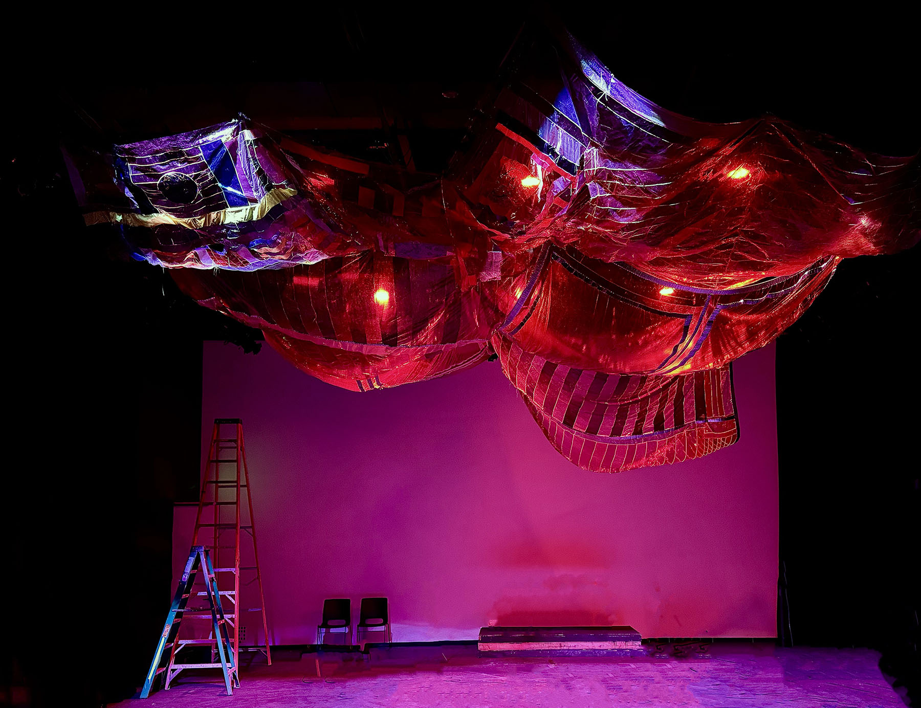 Stage with pink backdrop, colorful hanging fabric decorations, ladder, and two chairs, illuminated by red and purple lights.